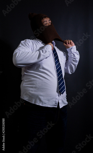 Obraz na plátně Adult Southeast Asian male wearing a brown knit balaclava mask, pulling down the lower edge of the disguise around the neck against a black background