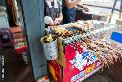 Foto Fresh whole squid on wooden skewers grilling over an open rack with sauce and a basting brush at a traditional Asian street food market stall