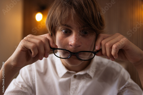 Konstfotografi Surprised young man wide-eyed and holding glasses, expressing shock and disbelie