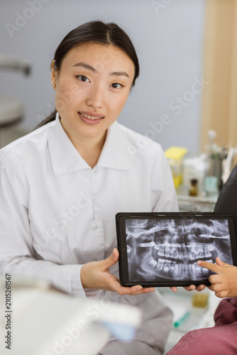 A female dentist in a white coat is showing a dental x-ray on a tablet to a patient, pointing at the screen