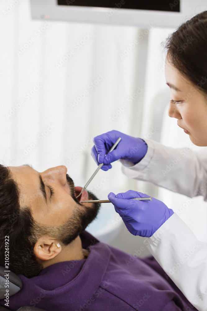 Fototapeta premium A female dentist in purple gloves and white coat performs a dental examination on a male patient lying in a chair