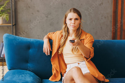 Young annoyed bored woman she 20s wear brown shirt casual clothes watch tv hold remote controller switch channels sit on blue sofa indoor rest at home in own room apartment. People lifestyle concept.
