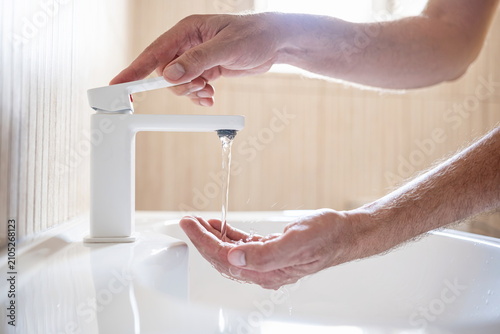 Close-up of a man turning on a white bathroom faucet while holding his other open hand under running water in a sink basin.