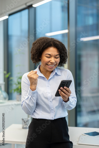 Vertical photo of an African American businesswoman standing outside an office building and rejoicing in success, looking at the mobile phone she is holding in her hand
