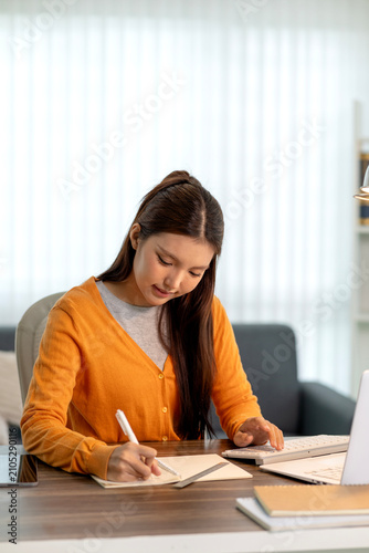 Young Asian Woman Writing Notes in Notebook While Studying Online at Home Office Desk