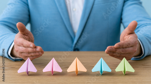Management and coaching leadership are essential for development and learning, illustrated by businessperson guiding colorful paper planes on wooden desk, symbolizing growth and strategy