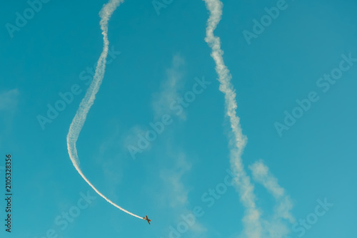 Portugal Oeiras Air Show. An airplane flies in the sky, making sharp turns and loops while creating smoke trails. The scene shows a bright blue sky with few clouds. The event occurs during daytime.