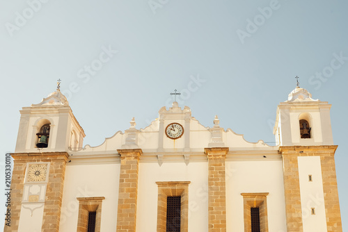 09 October 2023, Portugal, Evora - beautiful church stands tall with bell towers, clock on facade. bright sky above shows no signs clouds. scene captures the mid-afternoon light in a small town.