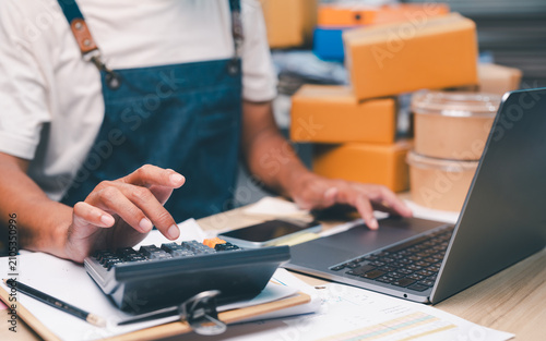 Person wearing an apron uses a calculator and laptop while managing paperwork and packages, likely handling business or inventory tasks.