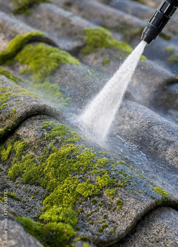 Person cleans moss from curved roof tiles using a pressure washer to remove green growth and restore surface. Action shows concentrated water jet removing moss and dirt.