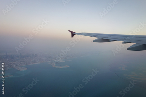 Aerial panoramic view of a modern coastal city captured from an airplane window during flight. The aircraft wing frames the skyline, with a large harbor, turquoise sea, shipping terminals, and dense 