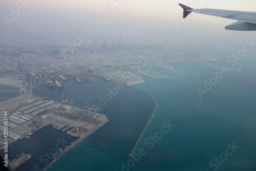 Aerial panoramic view of a modern coastal city captured from an airplane window during flight. The aircraft wing frames the skyline, with a large harbor, turquoise sea, shipping terminals, and dense 