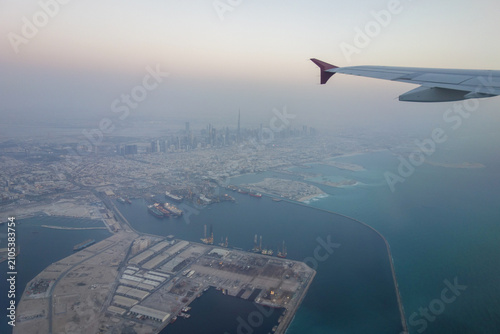 Aerial panoramic view of a modern coastal city captured from an airplane window during flight. The aircraft wing frames the skyline, with a large harbor, turquoise sea, shipping terminals, and dense 