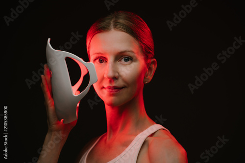 woman holding infrared light therapy mask and looking to the camera. black  background. Contrast red light. horizontal composition