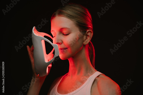 infrared light therapy mask. woman is holding the mask with closed eyes her face is illuminated by red light. black  background. Contrast red light. Horizontal composition
