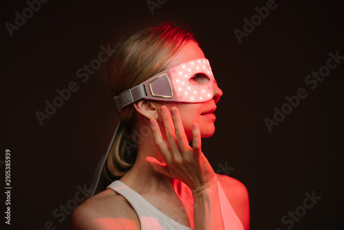 Red light therapy eye LED mask. woman profile wearing infrared light therapy mask. touching the mask with her hand.  looking side. dark background