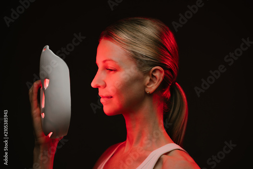 infrared light therapy mask. woman is looking at the mask her face is illuminated by red light. black  background. Contrast red light. Horizontal composition