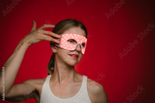 Red light therapy eye mask. smiling woman profile wearing infrared light therapy mask. touching the mask with her fit hand. dark background