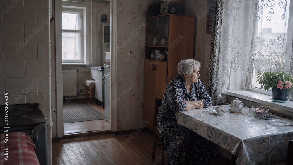Fototapeta premium Elderly woman sitting alone at a kitchen table by the window in a modest rural home