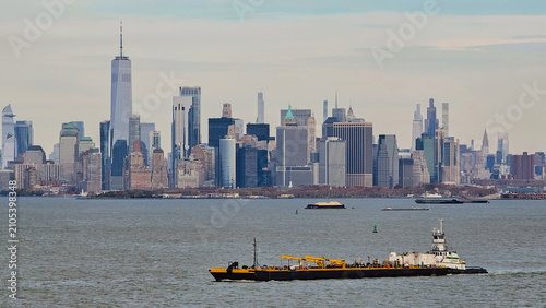 Panoramic view of Lower Manhattan skyline with Freedom Tower skyscrapers and an industrial tugboat barge vessel sailing across New York Bay on an overcast day