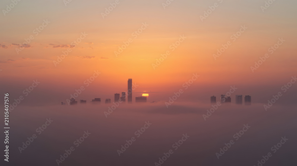 Fototapeta premium Misty sunrise skyline with distant skyscraper peeking above thin morning fog, serene light