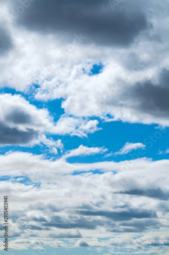 Picturesque blue sky texture, vertical vibrant cloudscape with a mix of fluffy white cumulus clouds, darker overcast storm clouds, and windows of bright blue sky