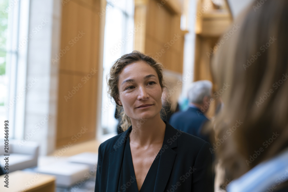 Fototapeta premium A well-dressed woman engages in conversation with a colleague in a bright, modern interior, highlighting professional interactions in contemporary workspaces.