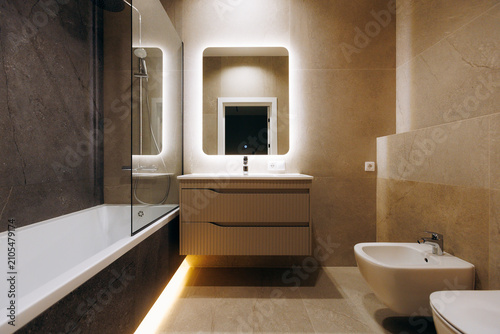 Modern bathroom interior with illuminated mirror, floating vanity, built-in bathtub, glass shower screen, wall-hung bidet, warm stone tile and soft LED lighting