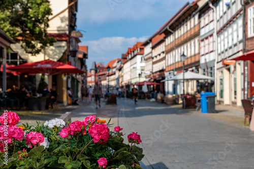 Photos Pink geranium flowers in focus in the foreground with blurred historic half-timbered houses along a pedestrian street in Wernigerode