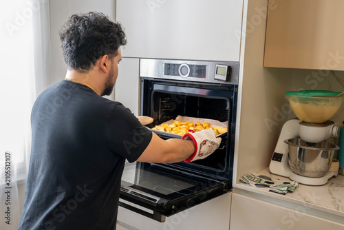 Rear view of a middle aged man cooking potatoes in a built in electric oven inside a bright modern residential kitchen