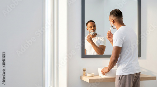 African American man shaving in bathroom mirror - Young black man shaving in modern bathroom while looking in mirror  