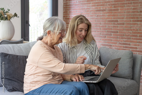 Young woman teaching mature adult senior lady how to use a laptop  two women sitting on a sofa at home  sharing online content  generational learning