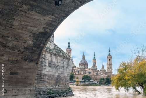 Autumn atmosphere in Zaragoza featuring the Basilica of Our Lady of the Pillar beyond a massive bridge pillar. Yellow trees and the turbulent Ebro River reflect a moody day in Spain