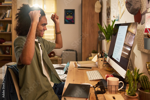 Konstfotografi Young adult man celebrating at desktop computer while reading acceptance message in home workspace, expressing success and achievement