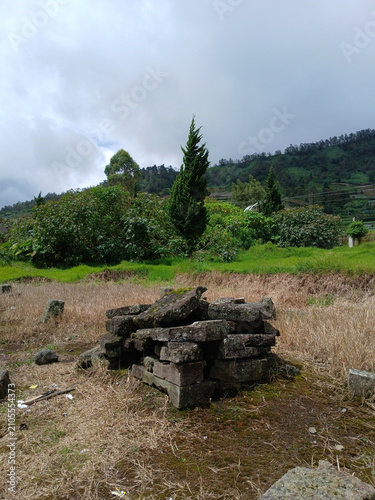 Ancient weathered stone blocks stacked neatly in a rural field with lush green trees and misty mountain slopes under a cloudy sky captured in this high resolution natural outdoor photograph.