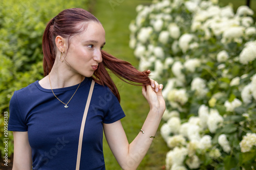 Fotografie Red-haired teenage girl touches her long hair while walking through blooming park on warm summer evening