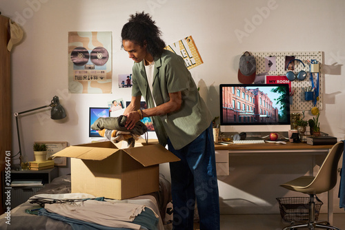Young adult man packing folded clothes into moving box in bedroom, preparing relocation or college move. Image supporting student housing, moving service, transition marketing