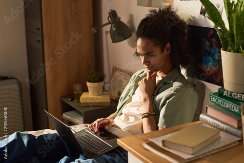 фотографія Young adult Black man studying on laptop in bedroom, researching coursework and thinking critically