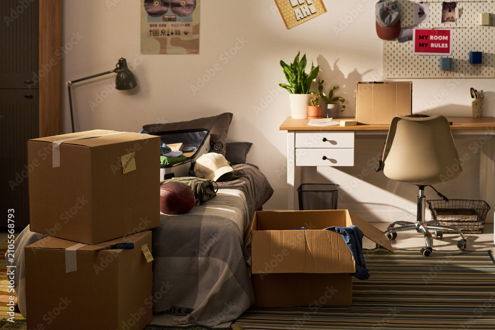 Naklejka premium Wide shot of teenager bedroom showing cardboard boxes, packed suitcase, desk, bed, signaling move in or move out. Supporting relocation, student housing, dorm life, transition marketing