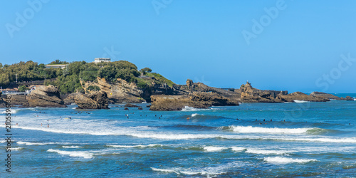 Crashing Atlantic Ocean waves breaking over dramatic black rock formations along the coastline in Biarritz under a brilliant blue sky with surfers in the water