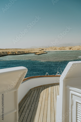 Yacht deck overlooking clear blue water and barren desert coastline under a bright sky
