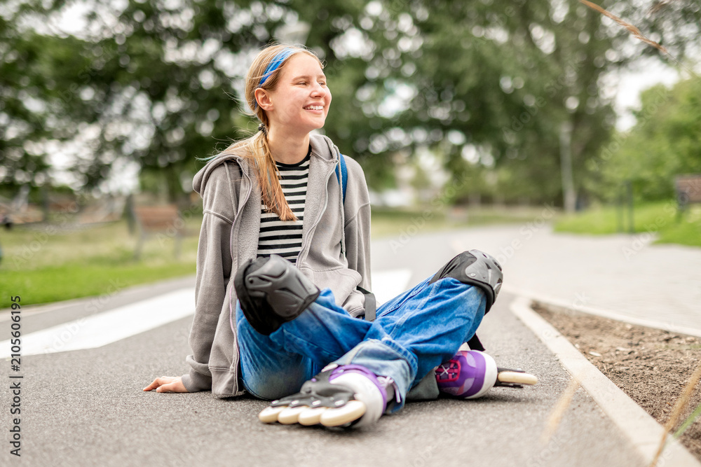 Fototapeta premium Blonde girl rests on park path. Wears rollerblades, protective gear.