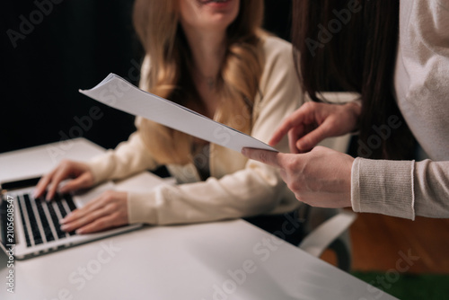 Cropped shot of two businesswomen collaborating on project, one typing on laptop while other provides feedback and advice on documents, representing mentorship and teamwork in office setting.