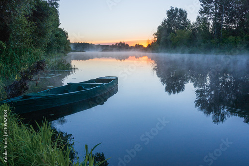 A serene wooden boat rests near the grassy riverbank during a misty sunrise, reflecting beautifully on the calm water surface