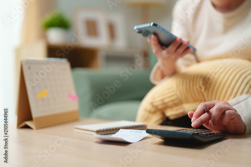 Принти на полотні Woman using smartphone and calculator to manage household budget with receipts, notebook and calendar at home