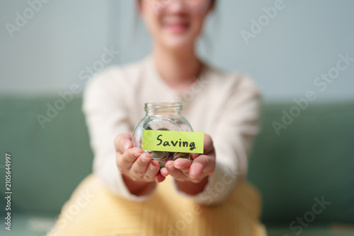 Fotografi Smiling woman holding a glass savings jar with coins at home, personal finance,