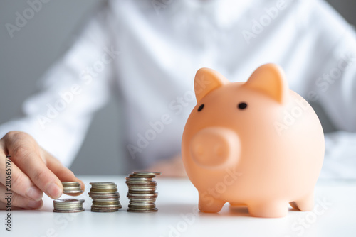 Coins are stacked next to a piggy bank while someone adds more coins to the pile during a money saving session