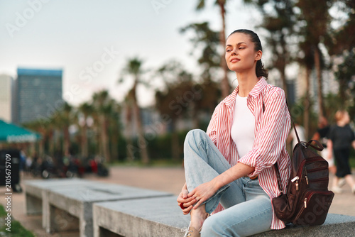 Принти на полотні A young woman student sits on a concrete ledge outdoors with a backpack, enjoying a quiet moment of study and reflection, sunlight warming her relaxed pose