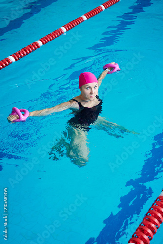Young woman swimmer in pool performing aqua fitness with dumbbells, arms extended in water, wearing swimsuit and cap, resistance training and aquatic exercise concept