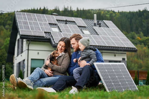 Family with child and dog sitting on grass near house with solar panels. Parents enjoying time together, caring for pet, sustainable lifestyle and renewable energy concept
